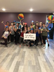 A group photo of people in workout clothes and costume accessories surrounding a sign that reads "Cast all your votes for dancing"